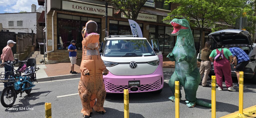 Pink and white VW EV with 2 people dressed as dinosaurs